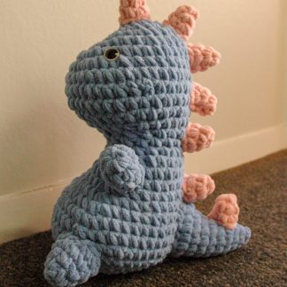 Blue crocheted plush dinosaur with blue and black eyes and pink stripes down the back