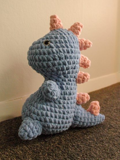 Blue crocheted plush dinosaur with blue and black eyes and pink stripes down the back